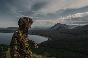 a young man in a khaki jacket stands with his back, arms outstretched, mountains