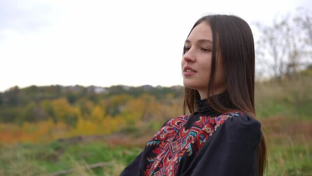 Zoom in to confident smiling young woman standing outdoors on overcast day admiring autumn river. Happy charming Ukrainian lady in black embroidered dress dreaming enjoying leisure