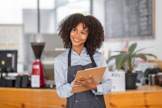 Female African coffee shop small business owner wearing apron standing in front of counter performing stock check. afro hair employee Barista entrepreneur.