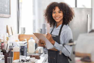 Female African coffee shop small business owner wearing apron standing in front of counter performing stock check. afro hair employee Barista entrepreneur.