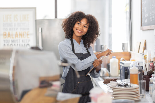 Female African coffee shop small business owner wearing apron standing in front of counter performing stock check. afro hair employee Barista entrepreneur.