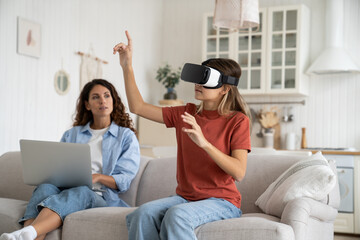 Interested teenage girl uses VR headset to visit metaverse or watch 3D virtual reality movies from comfort home. Progressive European schoolgirl waving hands sits on sofa near mother woman with laptop