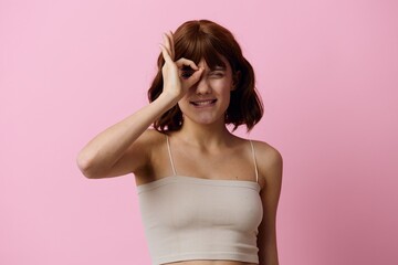 a funny, playful woman with short hair poses standing on a pink background with her fingers showing the OK sign near her eye, and her mouth wide open