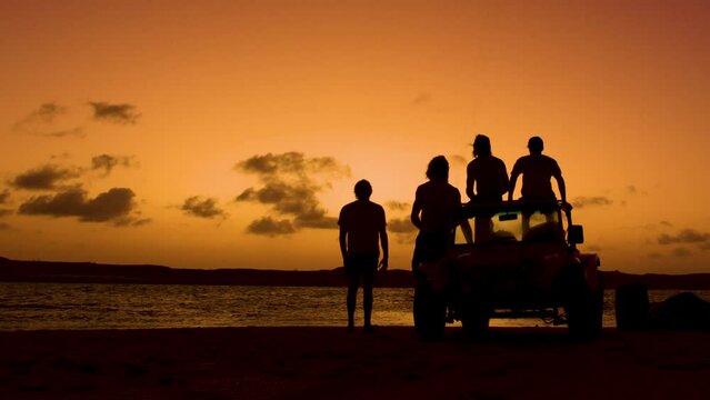 Silhouette showing group of people with quad watching sunset and jumping kiteboarder in ocean - static wide shot
