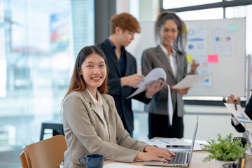 Happy beautiful Asian businesswoman sitting smiling and enjoying working on laptop in office and looking at camera.