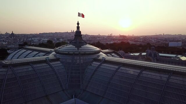 Aerial Tour of Paris city from above, featuring French flag waving at the top of The Grand Palais at sunset, with Sacre Coeur at distance background.