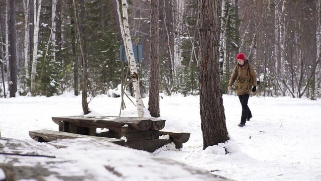 Man in a winter forest walking a large wooden table in a resting place and putting his travel backpack on a bench