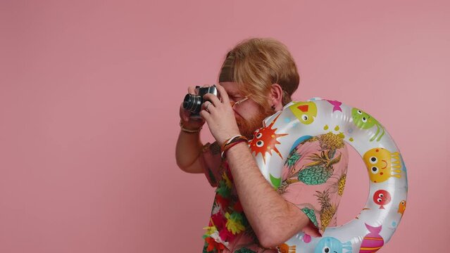 Happy hipster bearded man tourist photographer in sunglasses taking pictures photos on retro camera. Travel, summer holiday vacation. Hippie redhead smiling guy traveler on studio pink background