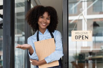 Small business african female owner smiling while turning sign for opening of cafe. Happy afro-american waitress entrepreneur in apron present sign on door