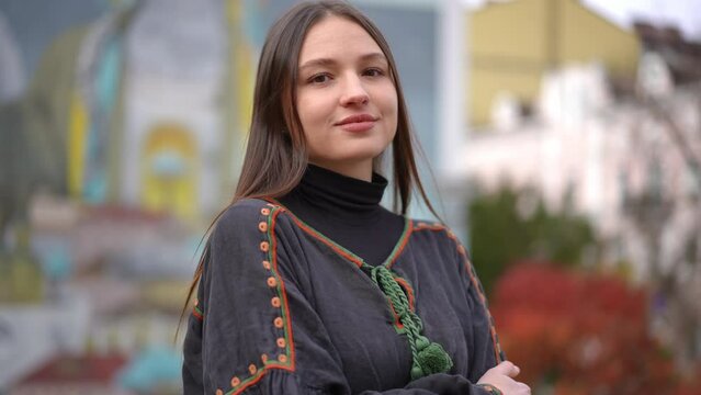 Young Ukrainian happy woman looking at camera smiling and looking around in slow motion. Portrait of brunette confident lady with brown eyes in embroidered shirt standing on city street