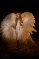 A girl in a white shirt, wings and with long hair, looking like a angel. A young model posing at a photo shoot with smoke, fog, flour, dust. A little fairy girl on a dark black background