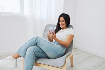 Woman sits on chair with phone in hand in new apartment, online shopping via phone, smile with teeth