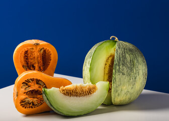 Two halves of an orange pumpkin and a slice of mountain melon on a white table.