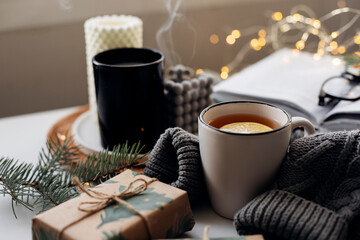 Winter home cozy concept. Mug with lemon tea, open book, warm sweater, candles and fir tree....