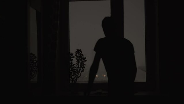Dark silhouette of slender young man approaches window of room and looks out into street. Night snow storm, snow sticks to the windows. Home plant and book are on the windowsill.