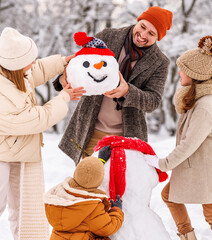 Happy children sculpting funny snowman together with parents in winter snow-covered park