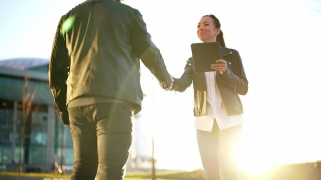 Man and woman stand on nature landscape in sun rays and shake hands as sign of greeting or respect. Girl holding tablet in hands. Business meeting two people. Back view on interlocutor and handshake.