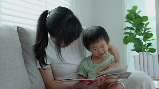 Happy Asian mother relax and read book with baby time together at home. parent sit on sofa with daughter and reading a story. learn development, childcare, laughing, education, storytelling, practice.