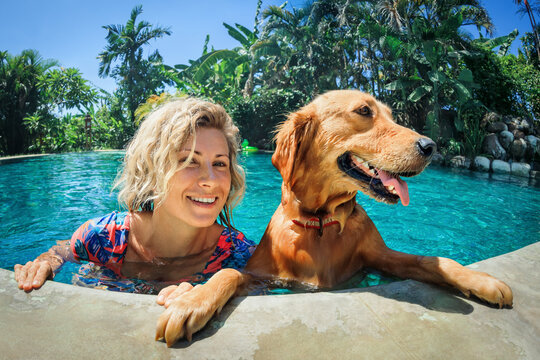 Funny portrait of smiling woman relaxing with golden retriever puppy in tropical swimming pool. Popular dog breeds, outdoor activity and fun games with family pet on summer family holiday.