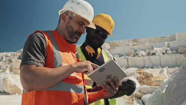 Medium shot of mature Caucasian foreman holding digital tablet standing in front of marble stone discussing work plans with African American miner