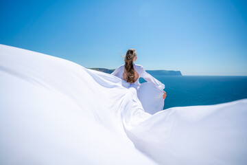 Fototapeta na wymiar Blonde with long hair on a sunny seashore in a white flowing dress, rear view, silk fabric waving in the wind. Against the backdrop of the blue sky and mountains on the seashore.