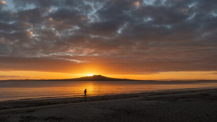 Fototapeta na wymiar Silhouette woman walking on Milford beach. Sun rising over Rangitoto Island. Auckland.