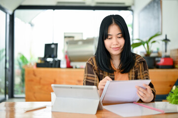 Fototapeta premium Charming Asian female remote working in the coffee shop, analyzing something on paperwork.
