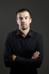 Fototapeta premium Portrait of man in a black shirt on a dark background. Serious brunet with arms folded. Young caucasian man