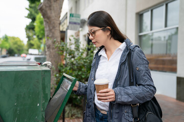 asian chinese female business person carrying coffee and opening green newspaper rack to check if thereâs still free tabloid to get on her way to work in the morning in palo alto city California usa - obrazy, fototapety, plakaty