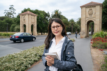 happy asian Taiwanese female exchange student holding book and smiling at camera on palm tree boulevard near entrance gate of school university in California usa