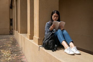 leisure asian korean girl college student sitting against pillar with bending kneels on the hallway and enjoying reading a book on school campus in California usa