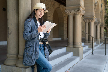 smiling asian Korean female visitor leaning against column from travel book while learning about Romanesque architecture on school campus in California usa