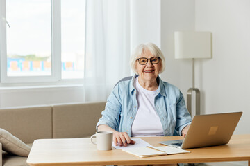 a stylish, successful elderly woman with gray hair works from home on a laptop and looks at the camera with a pleasant smile