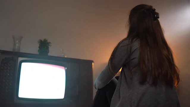 Broken vintage TV in dark illuminated room, woman sitting on the floor and watching television channels with bad cable signal and blinking stripes on the screen. Retro TV entertainment at home