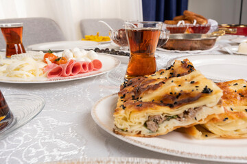 Turkish patty, close up photo of Turkish patty called su borek with meat. Traditional breakfast table setup. Glass of fresh hot tea. Cheese plate, ham, salami, sausage called sucuk, bagel called simit