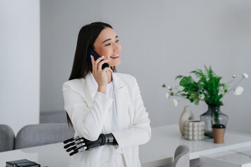 Cheerful gorgeous brunette girl with bionic arm talks by phone, smiles looks aside at home. Toothy...