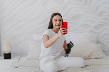 Attractive caucasian girl with bionic arm in white clothes  sits on bed makes video call by phone,...
