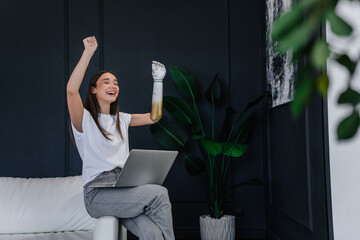 Cute brunette Caucasian girl with artificial hand sits on leather couch at home with Laptop raises...