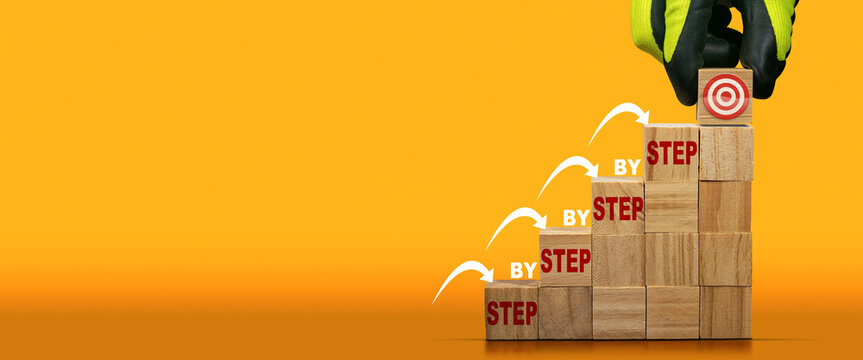 Gloved hand arranging wood blocks as step stair with a target symbol on the last step and text Step by Step. Ladder of success or goal concept. Orange and yellow background with copy space.