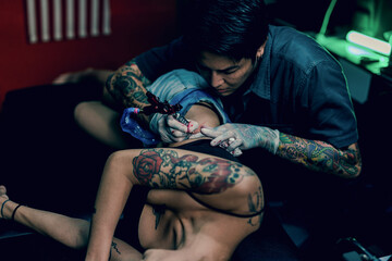 Professional tattooist makes the tattoo on a girl waist, focusing on tattoo machines in a modern studio lowlight.