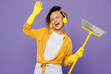 Young housekeeper woman wear yellow shirt rubber gloves headphones listen music dance tidy up hold...
