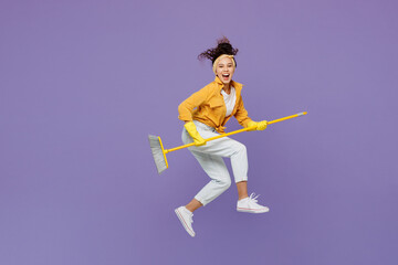 Fototapeta na wymiar Full body side view smiling young housekeeper woman wear yellow shirt tidy up jump high hold broom sweep floor look camera isolated on plain pastel light purple background studio. Housework concept.