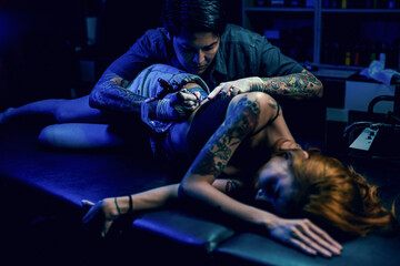 Professional tattooist makes the tattoo on a girl waist, focusing on tattoo machines in a modern studio lowlight.