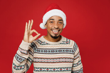 Merry young smiling cool man wear warm cozy Christmas sweater Santa hat posing showing okay ok gesture isolated on plain red color background studio. Happy New Year 2023 celebration holiday concept.