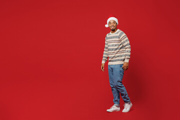 Fototapeta na wymiar Full body side view smiling fun merry young man wear warm cozy Christmas sweater Santa hat posing walk go look camera isolated on plain red background. Happy New Year 2023 celebration holiday concept.