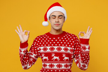 Merry young man wear red knitted sweater Santa hat posing hold spread hand in yoga om aum gesture...