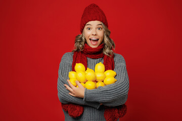 Young excited woman wear grey sweater scarf hat hold in hands bunch of lemons fruits isolated on plain red background studio portrait. Healthy lifestyle ill sick disease treatment cold season concept.