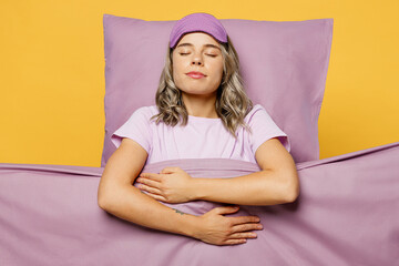 Young woman wear purple pyjamas jam sleep eye mask rest relax at home lies wrap covered with blanket duvet close eyes hold belly isolated on plain yellow background studio Good mood night nap concept