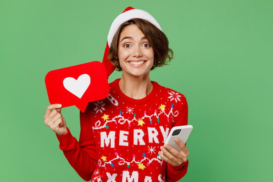 Merry young woman wears xmas sweater Santa hat posing hold in hand use mobile cell phone heart form like icon sign isolated on plain pastel light green background. Happy New Year 2023 holiday concept.