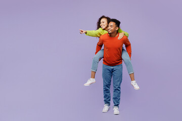 Full body young couple friends family man woman of African American ethnicity wear casual clothes together giving piggyback ride to joyful, sit on back point aside isolated on plain purple background.